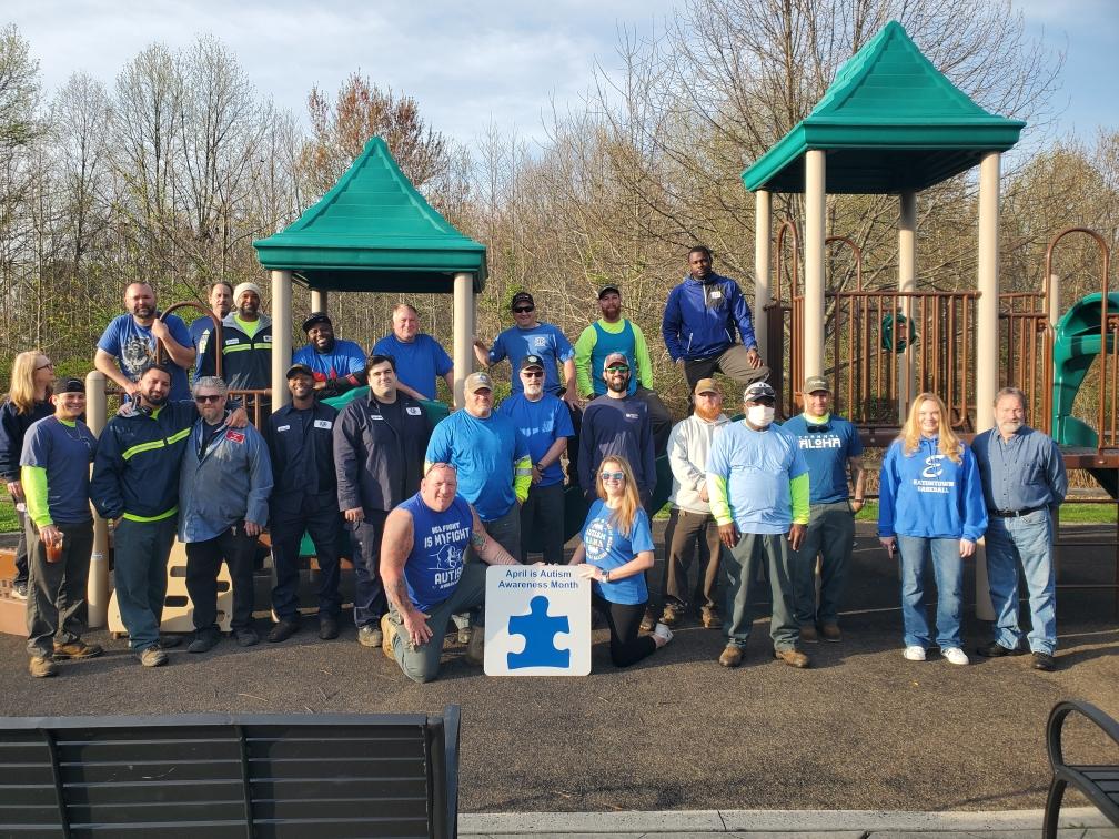 Group of adults posing outdoors at a playground; two green gazebo structures and an Autism Awareness Month sign.